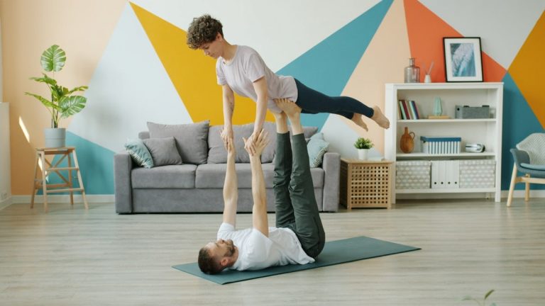 Happy couple is doing pair yoga at home, man is holding woman with legs on floor, girl is stretching arms. Relationship, active people and sports concept.